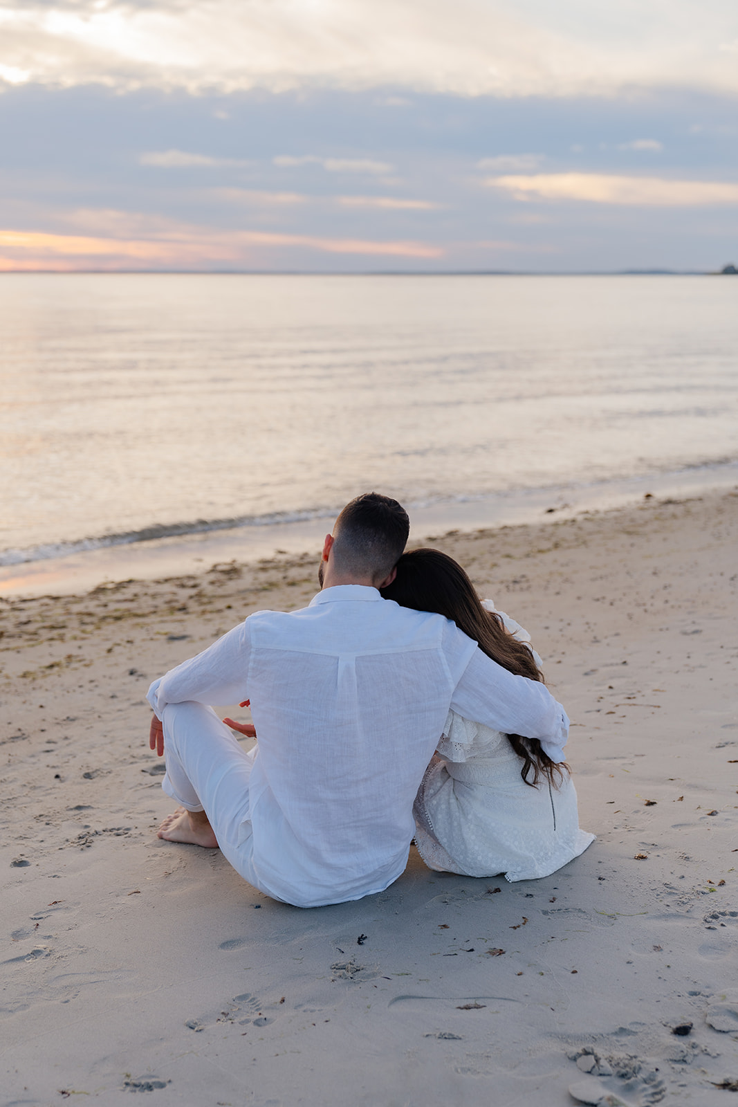 Cape Cod Engagement Photos of a couple sitting together on the beach, cuddled up with their backs facing the water as the sunset paints the sky.