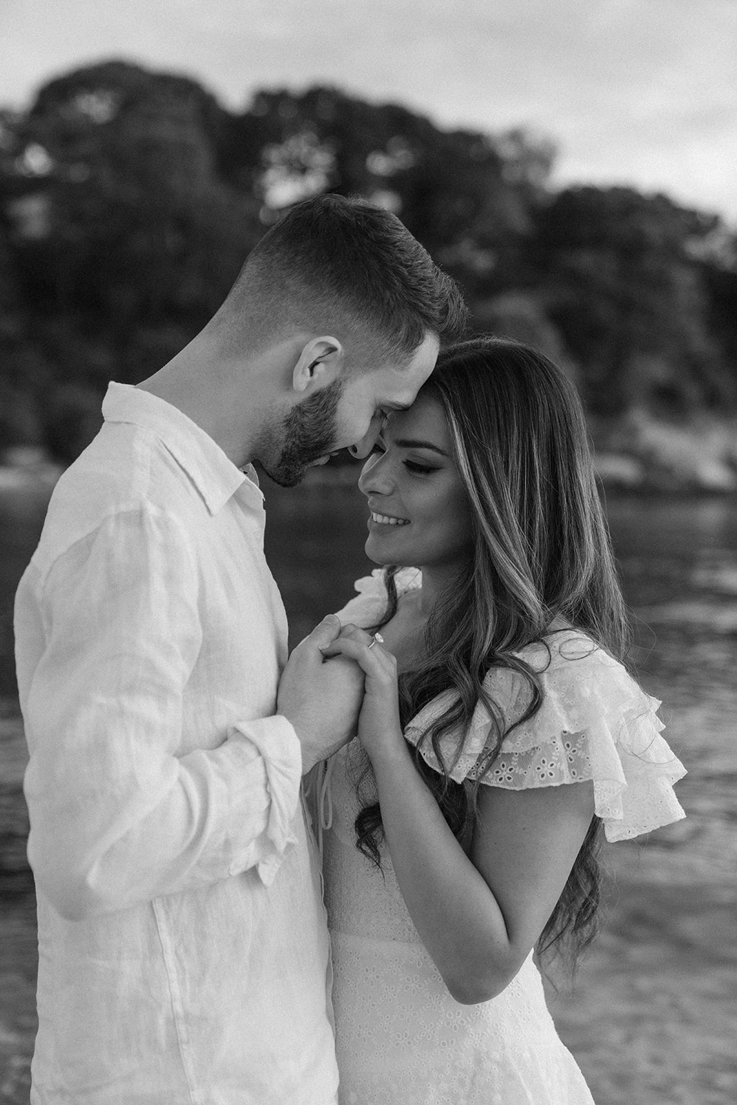 A black-and-white image of a couple smiling and gazing into each other's eyes, hands gently clasped as they share an intimate moment by the water.