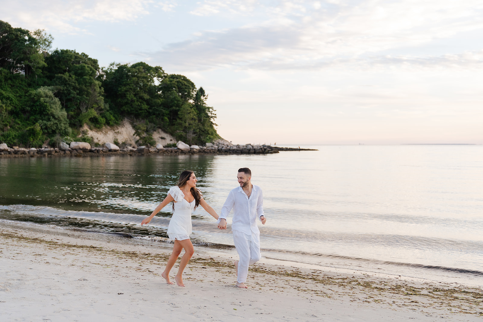 Cape Cod Engagement Photos of a couple running barefoot along the beach, laughing and enjoying a playful moment as the water gently laps the shore.