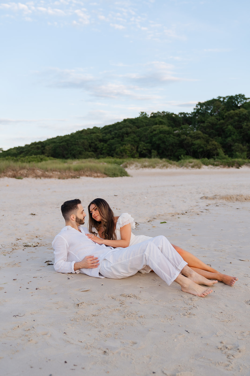 A couple lying on the beach, gazing at each other with smiles, surrounded by soft sand and the calm sea in the background.