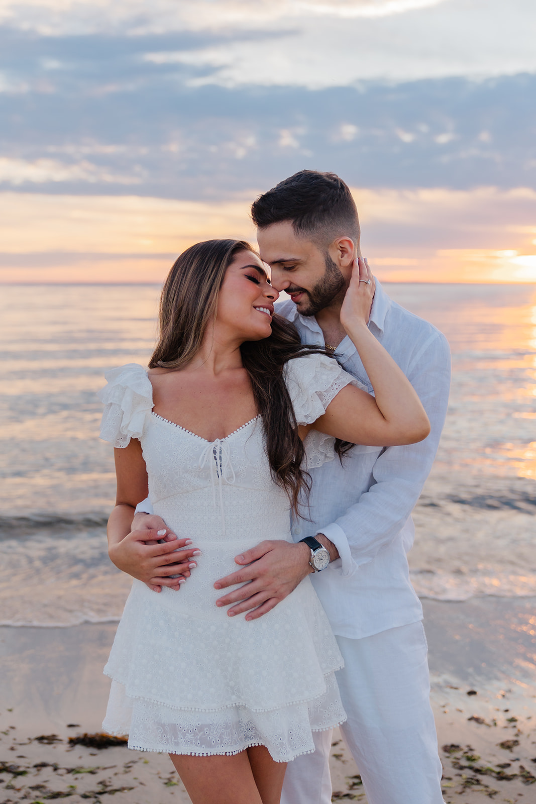 A couple embracing on the rocks near the water, sharing a kiss, with the soft reflection of the sky on the calm water in the background.