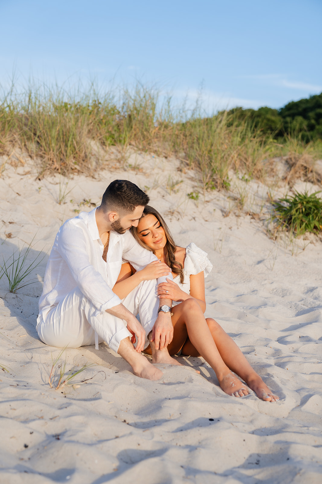 The couple sitting close together on the beach, with a peaceful backdrop of the Cape Cod coastline, capturing the sweet and intimate moment in their engagement photos.