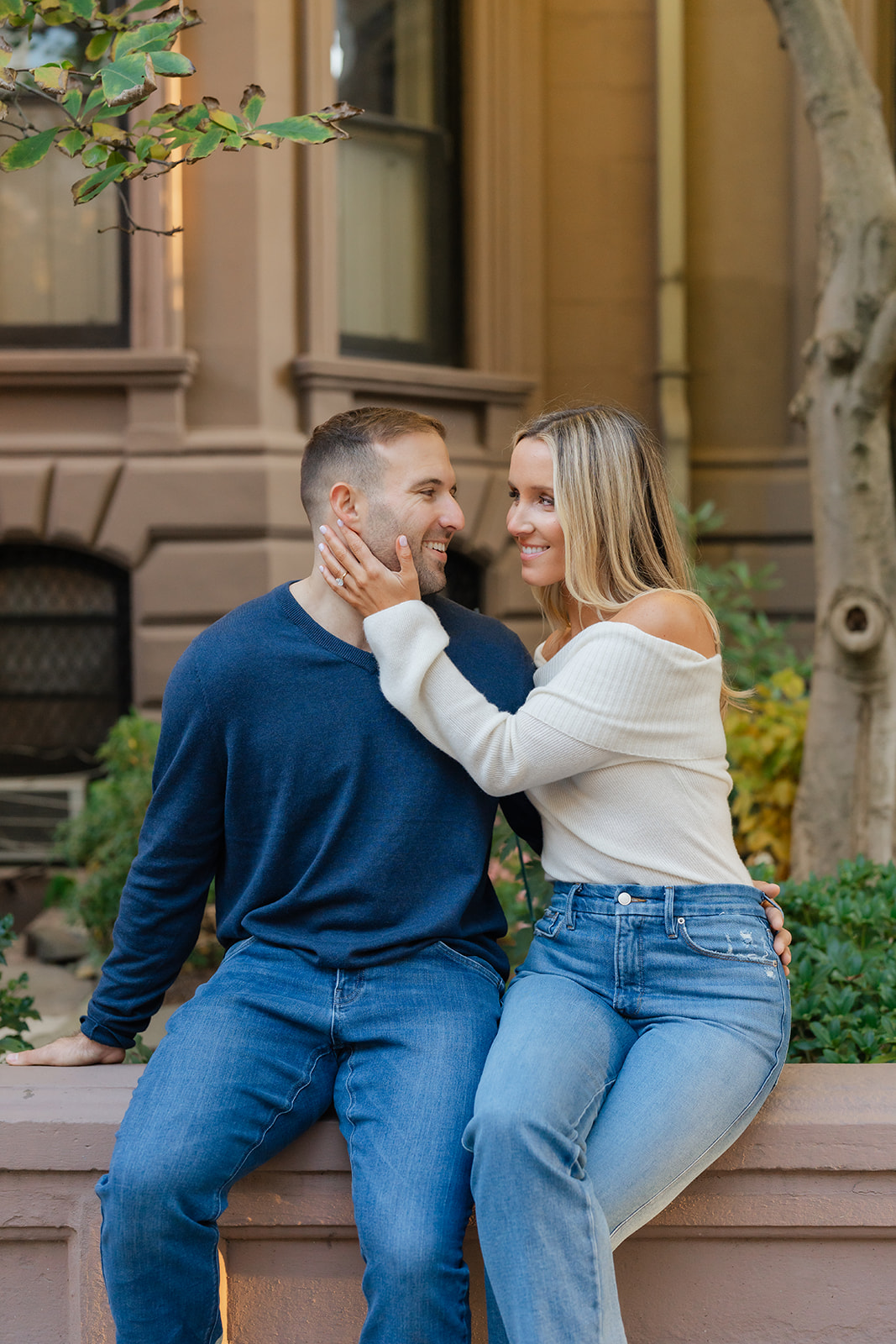 A couple shares a tender moment, with the man smiling and the woman playfully touching his cheek. This intimate photo captures a loving moment during their Boston Public Garden engagement photoshoot, showcasing the couple's connection against a neutral background of soft architectural details.