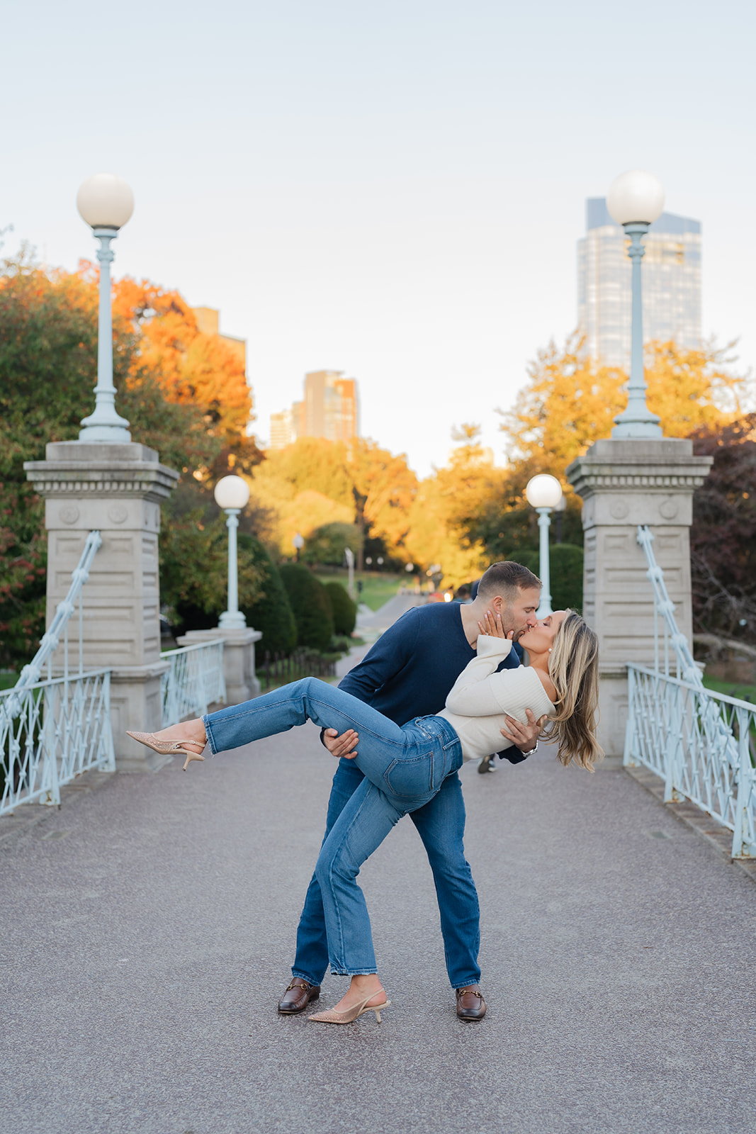 In this stunning Boston Public Garden engagement photo, a man lifts his fiancée into a romantic dip while they kiss in the golden hour sunlight. The historic bridge in the background and the autumn trees add to the romantic ambiance.