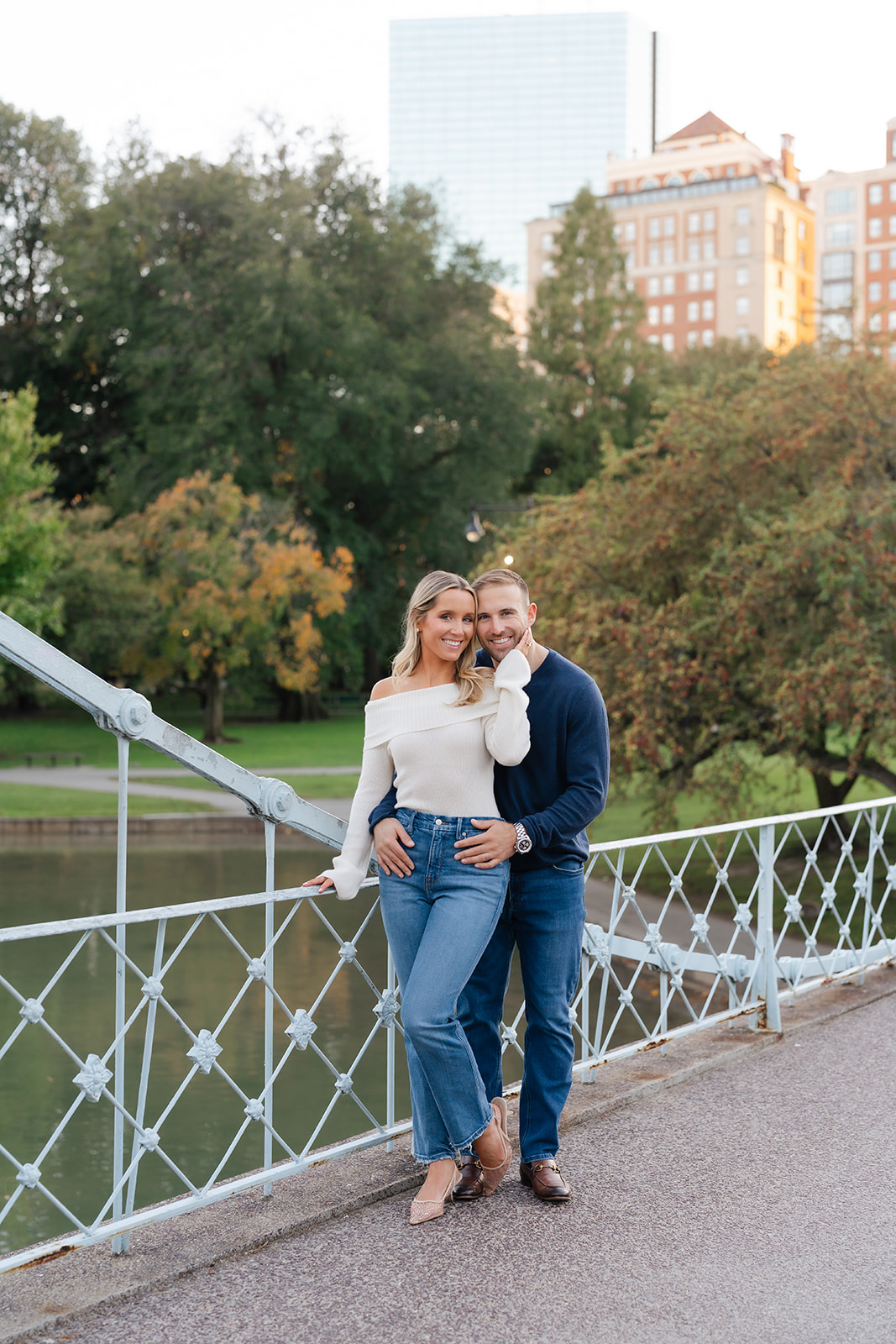 A couple poses for an intimate Boston Public Garden engagement photo as they stand close to each other on the bridge. The woman, dressed in an off-shoulder sweater and jeans, leans into her fiancé, smiling lovingly as they gaze at each other, with the city skyline in the background.