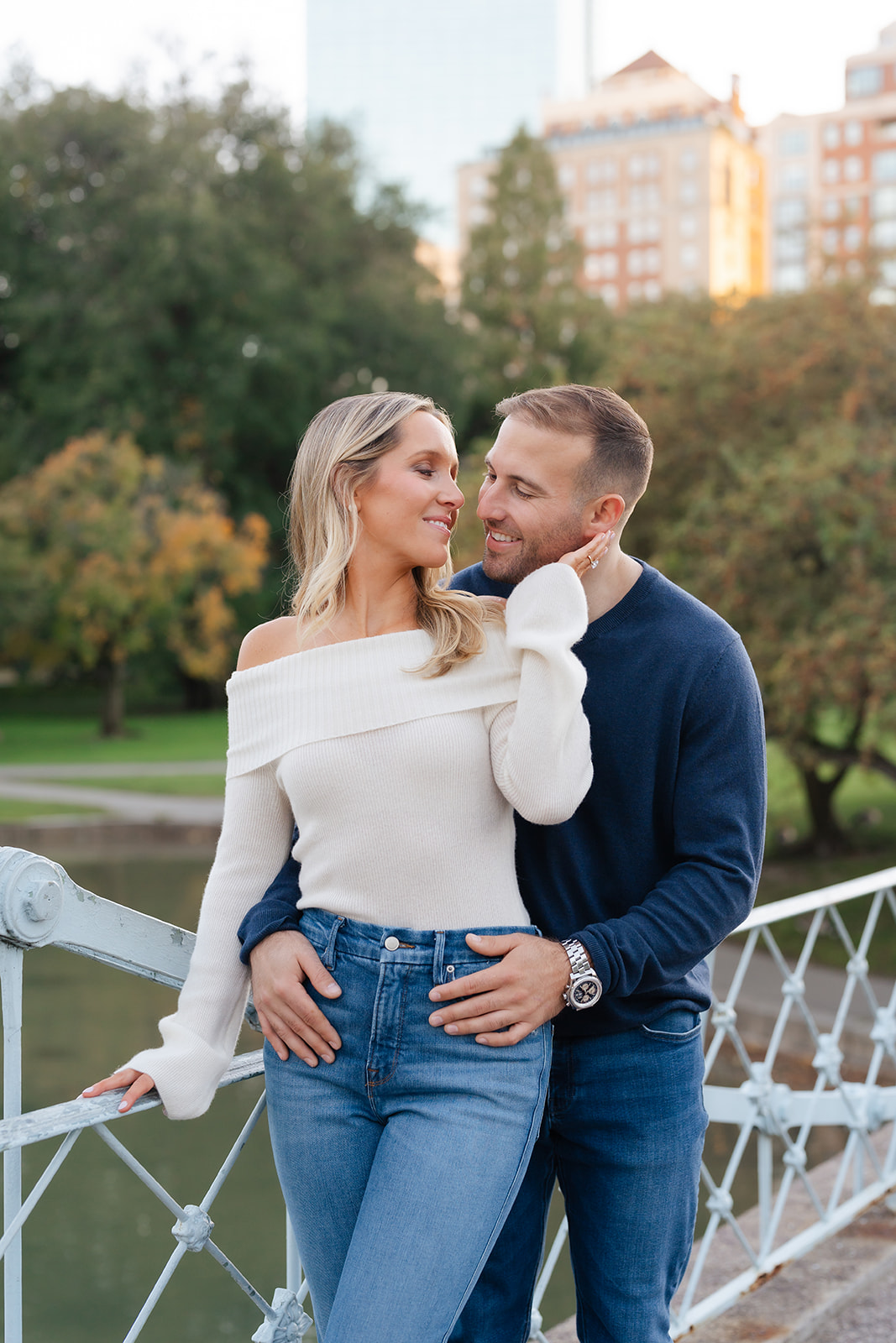 A couple shares a tender moment, with the man smiling and the woman playfully touching his cheek. This intimate photo captures a loving moment during their Boston Public Garden engagement photoshoot, showcasing the couple's connection against a neutral background of soft architectural details.