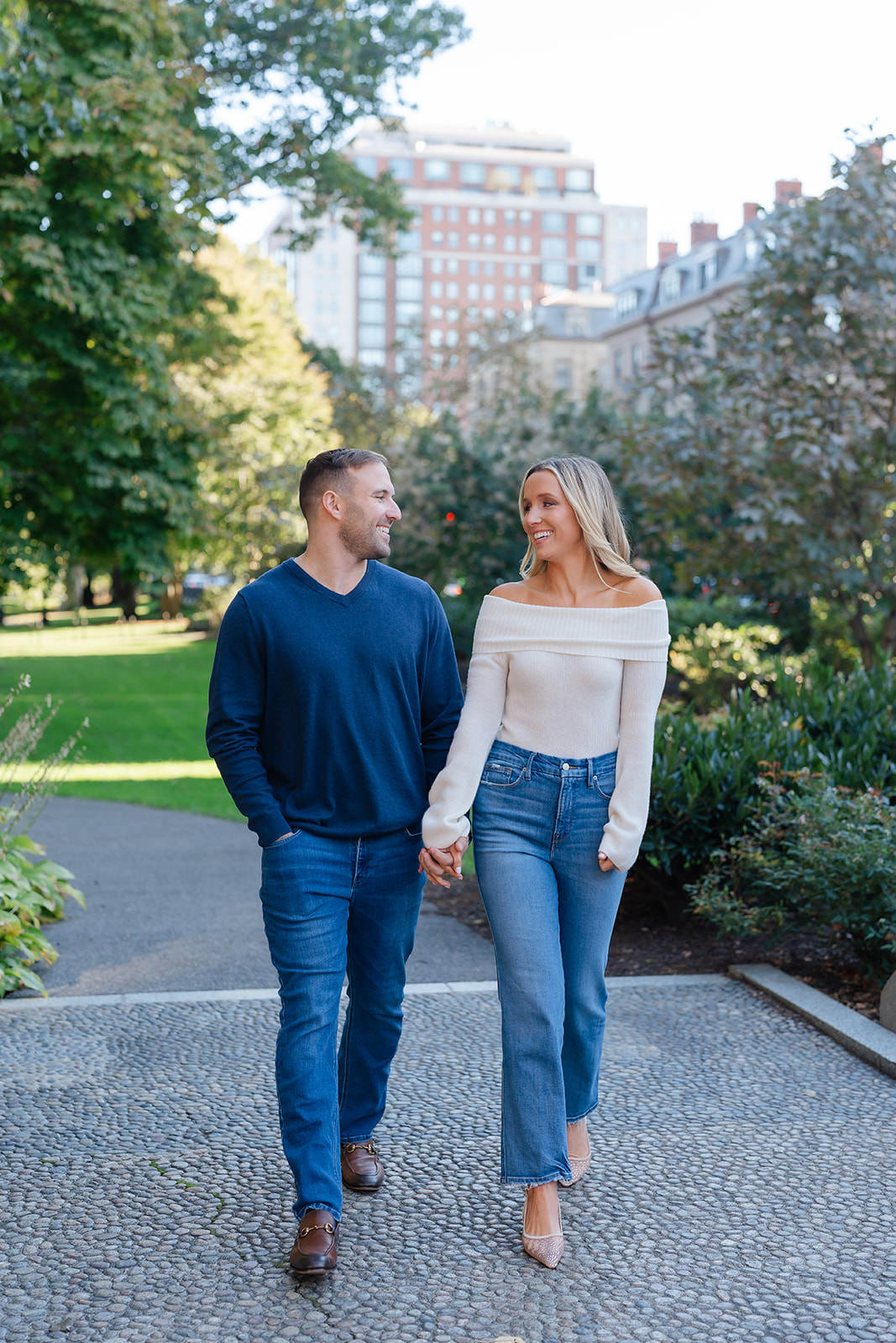 Couple walking hand-in-hand through the Boston Public Garden with a lush green background. Ideal for Boston Public Garden engagement photos.