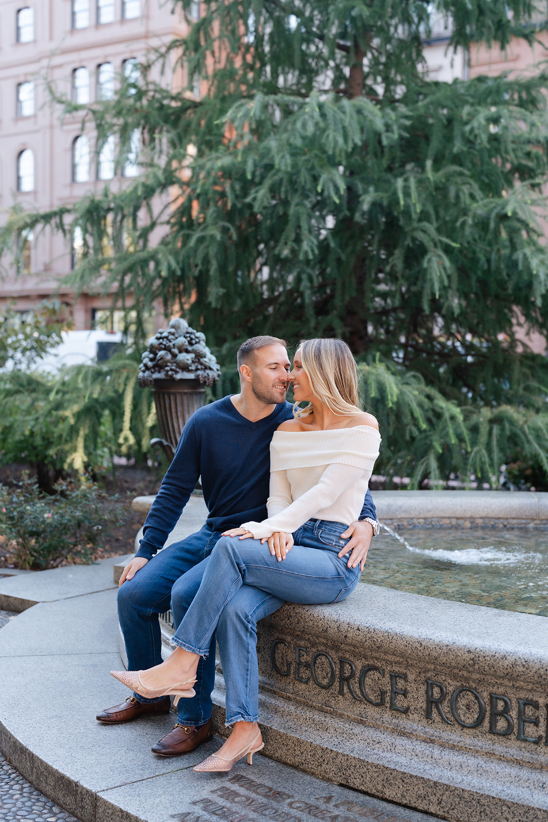 Couple sitting on a stone bench, sharing a sweet moment in the Boston Public Garden, with the iconic greenery and peaceful atmosphere surrounding them.