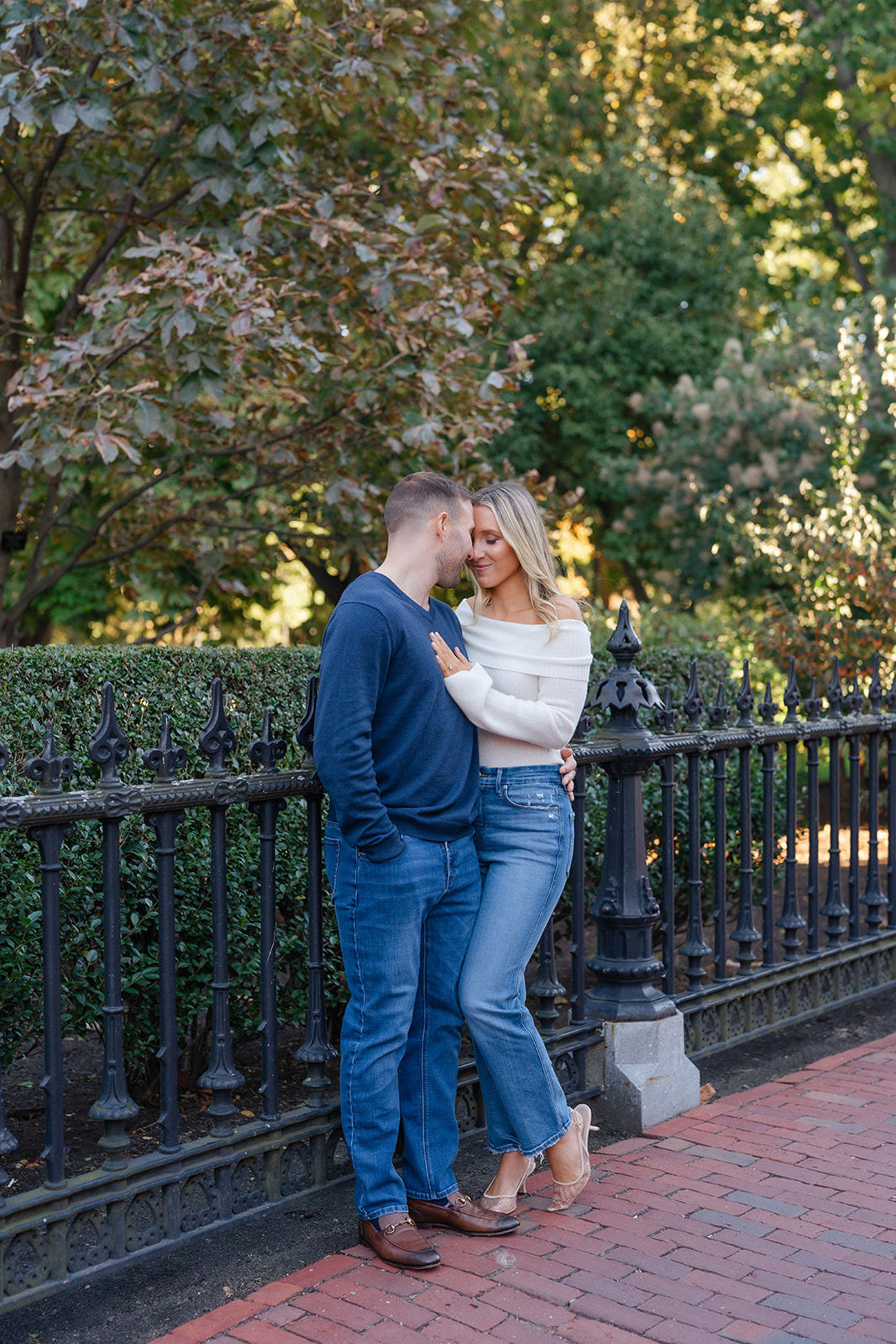 A couple in a loving embrace, with the woman gazing into her partner’s eyes while standing by a garden fence in the Boston Public Garden.