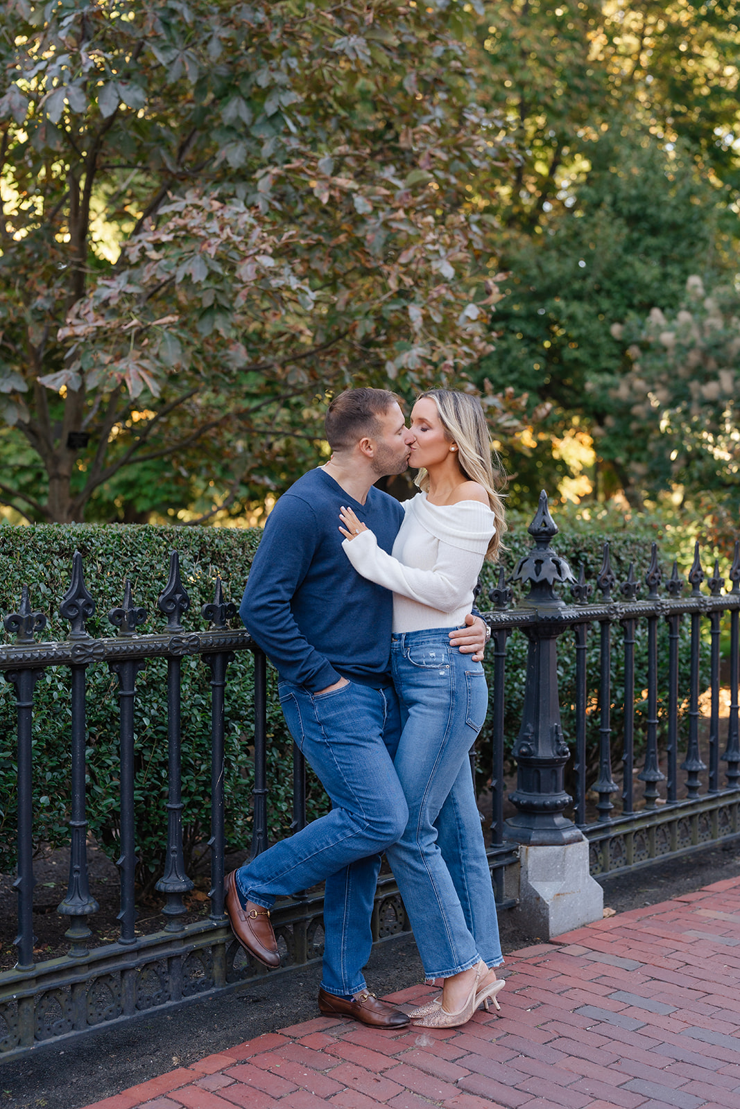 A couple in a loving embrace, with the woman gazing into her partner’s eyes while standing by a garden fence in the Boston Public Garden.