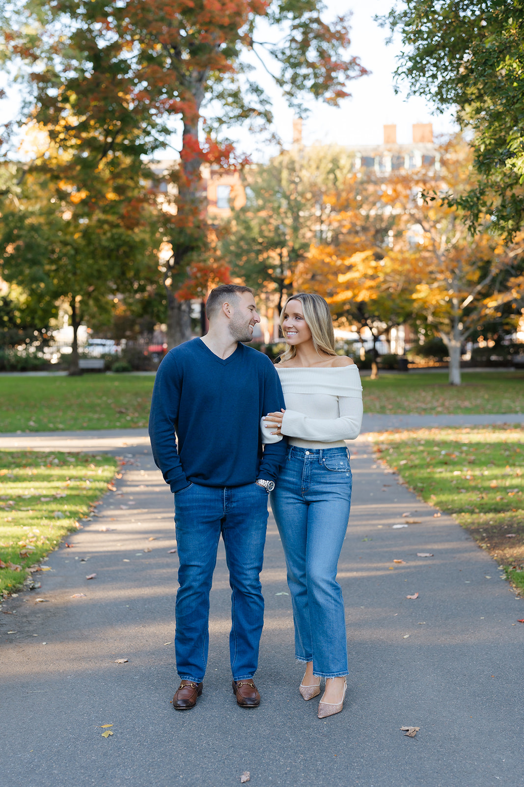 Couple laughing and holding hands while walking down a garden path in the Boston Public Garden, with beautiful autumn foliage in the background.