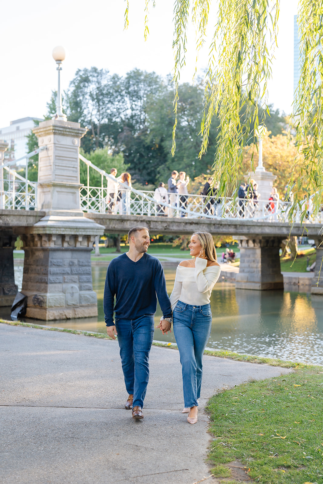 Couple walking hand-in-hand through the Boston Public Garden with a lush green background. Ideal for Boston Public Garden engagement photos.