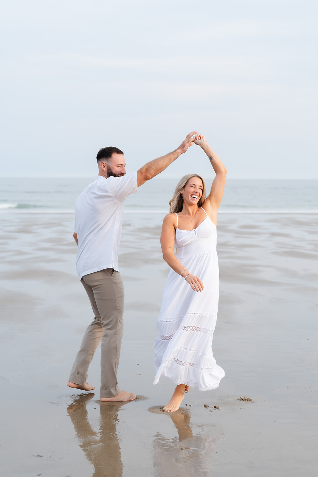 A couple twirling and dancing together barefoot on the beach, full of laughter and love during their Beach Engagement session.