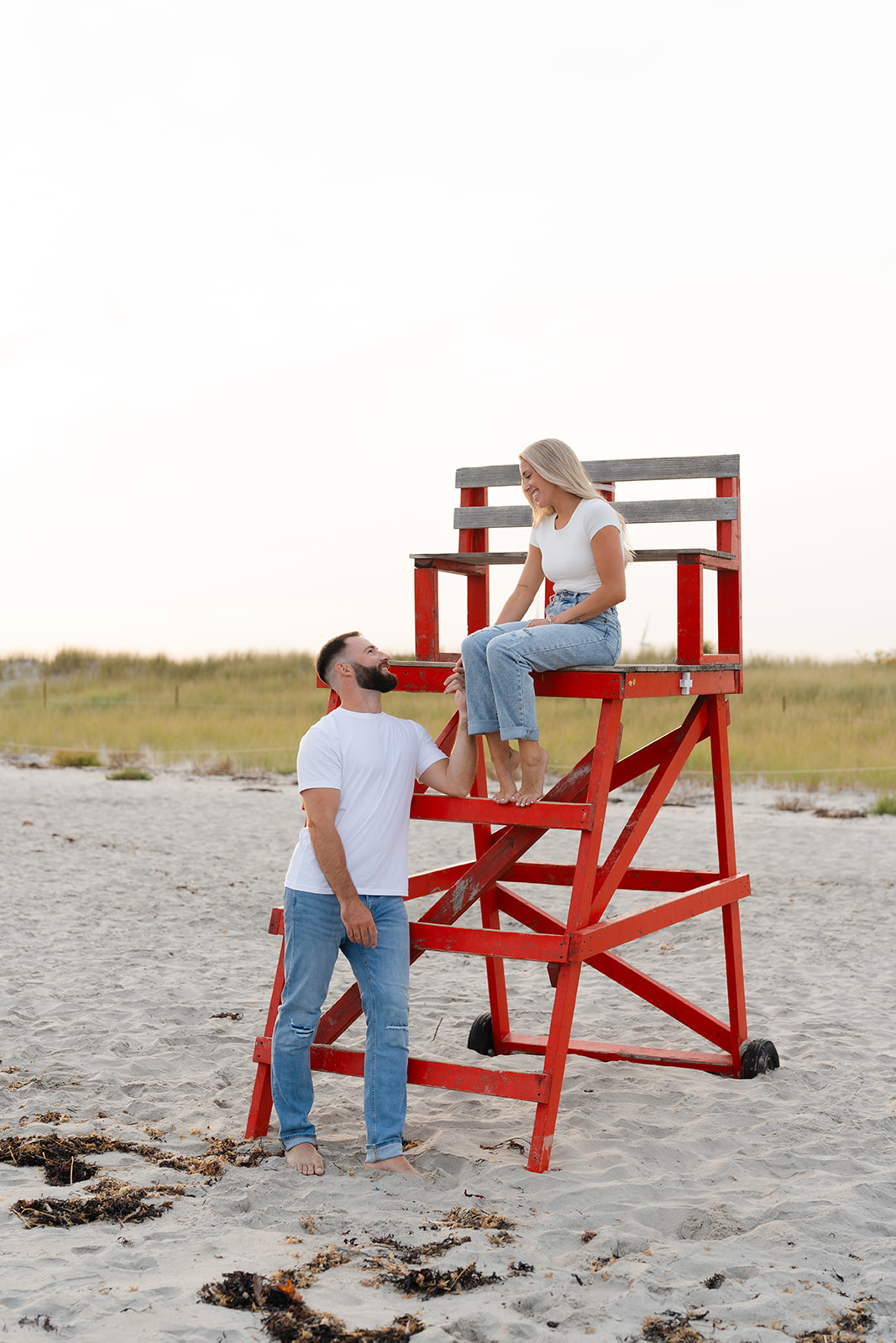 Couple posing for a playful moment, laughing while sitting on a lifeguard chair at the beach engagement session.