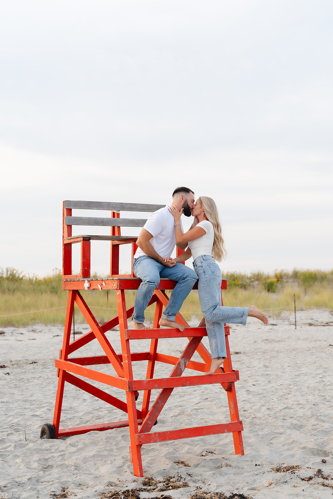 Couple posing for a playful moment, laughing while sitting on a lifeguard chair at the beach engagement session.