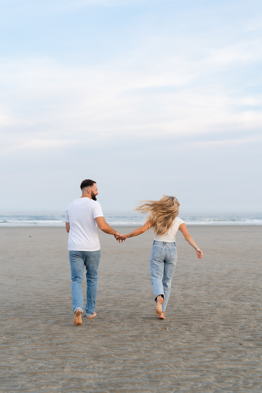 A playful couple running together on the beach during their engagement session, barefoot with the ocean in the background.