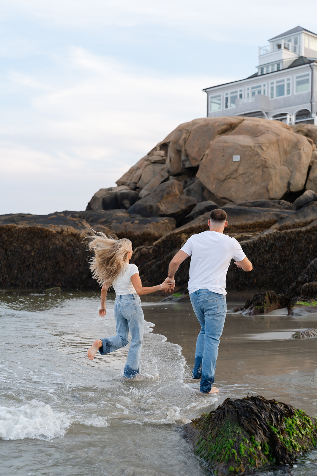 A playful couple running together on the beach during their engagement session, barefoot with the ocean in the background.