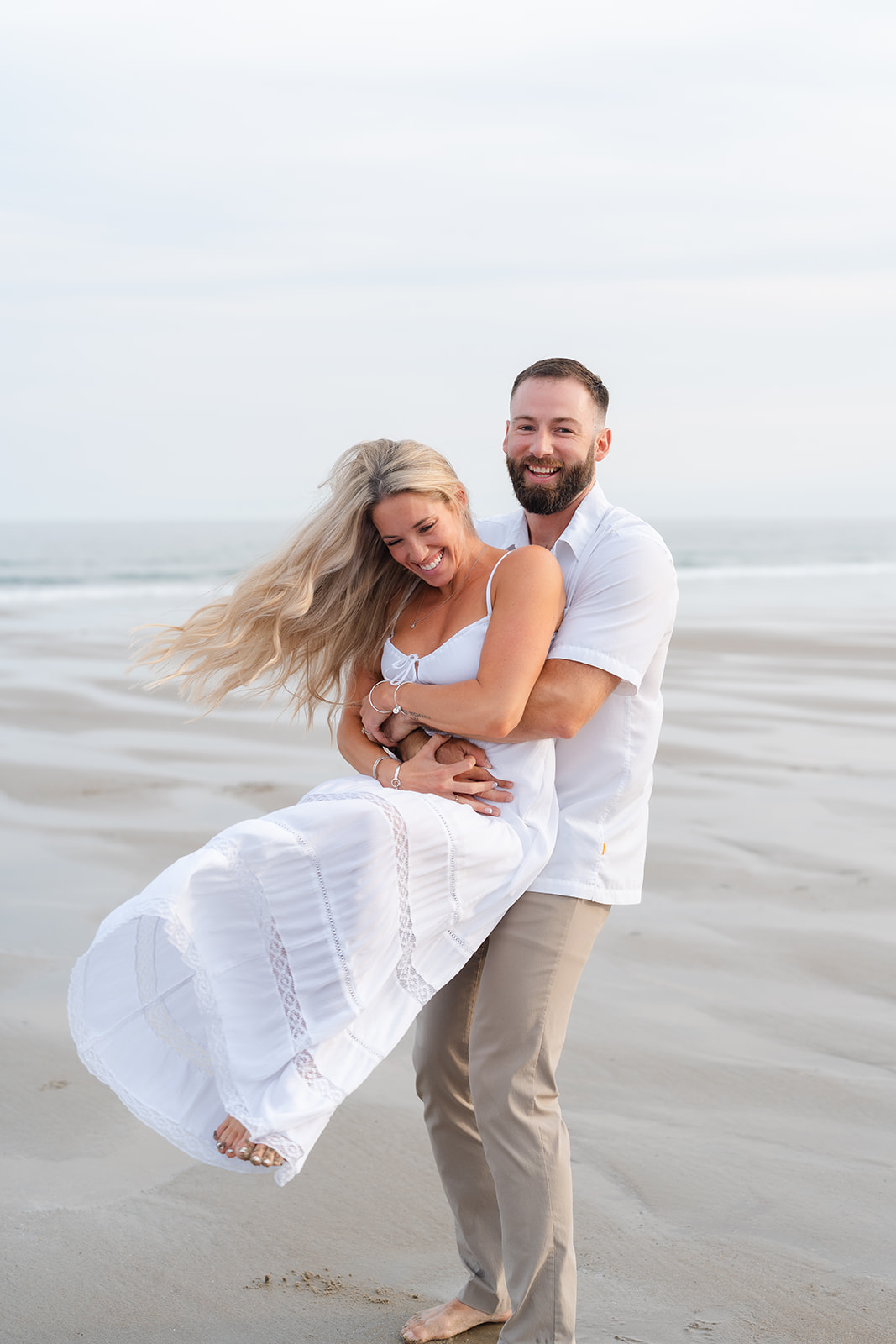 A couple laughing and playing, as one partner lifts the other off the ground, their joyful connection highlighted in this sweet Beach Engagement photo.