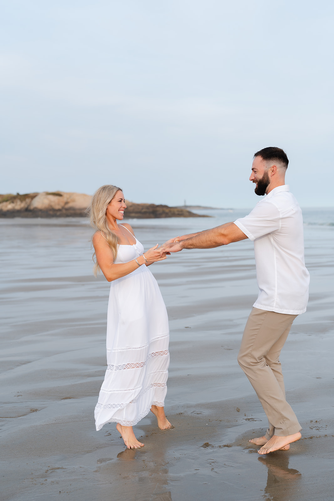 A couple holding hands and walking along the beach, with the water stretching out behind them, enjoying a candid moment during their Beach Engagement session.