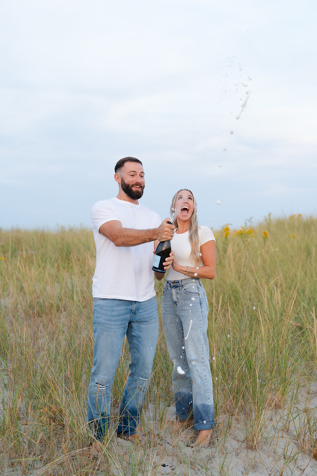 Couple holding champagne glasses and toasting during their beach engagement session, with their engagement ring clearly visible.