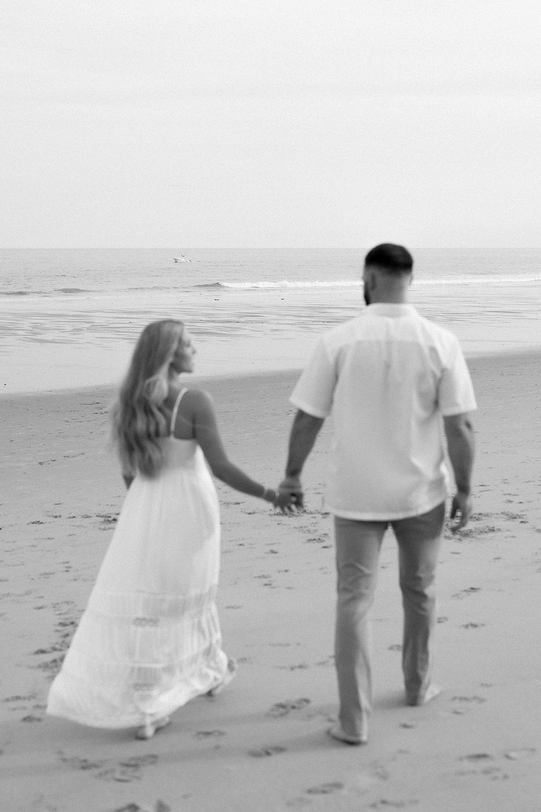 A black-and-white image of a couple holding hands as they walk along the beach, with the ocean in the background, capturing a serene moment during their Beach Engagement session.