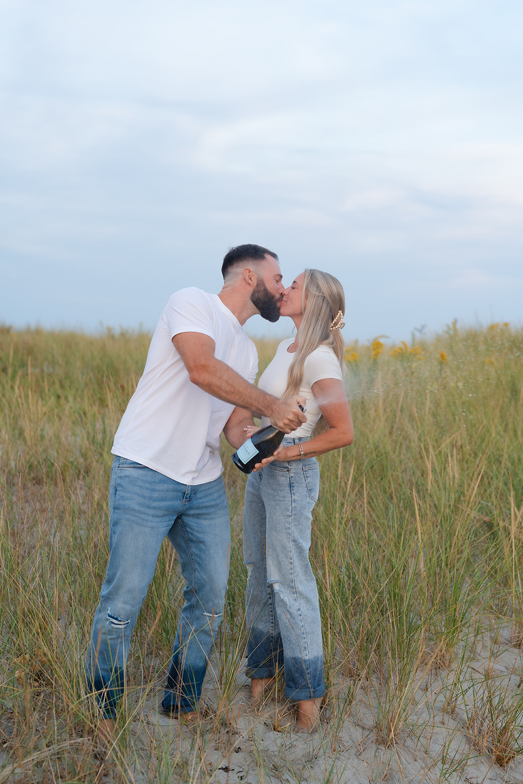 Couple holding champagne glasses and toasting during their beach engagement session, with their engagement ring clearly visible.