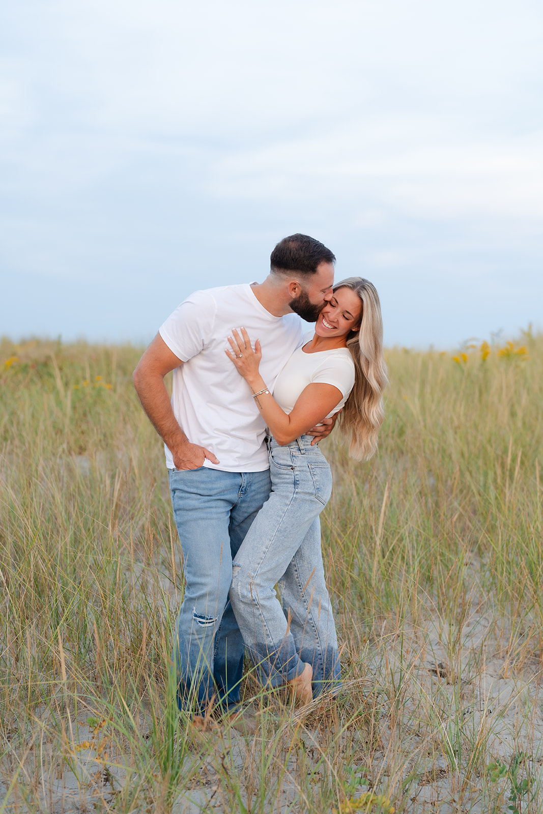 A couple cuddling and smiling as they embrace each other on the beach during their engagement session, showcasing genuine love and connection.