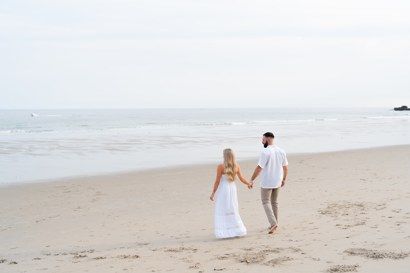 A joyful couple embracing each other in a playful moment during their Beach Engagement session on a sandy beach, dressed casually in white and beige attire.