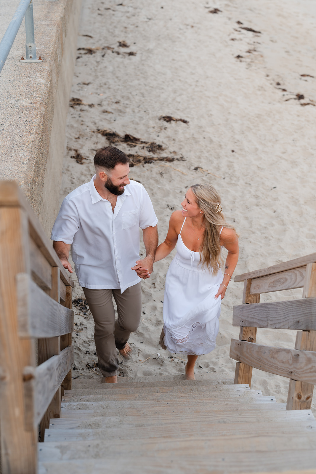 A couple walking up the beach stairs, holding hands and gazing at each other with smiles, capturing a fun and loving moment from their Beach Engagement session.