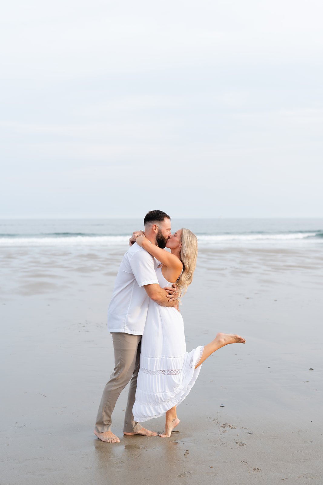 A couple sharing a passionate kiss on the beach, with one partner lifting the other, creating a romantic and fun moment during their Beach Engagement session.