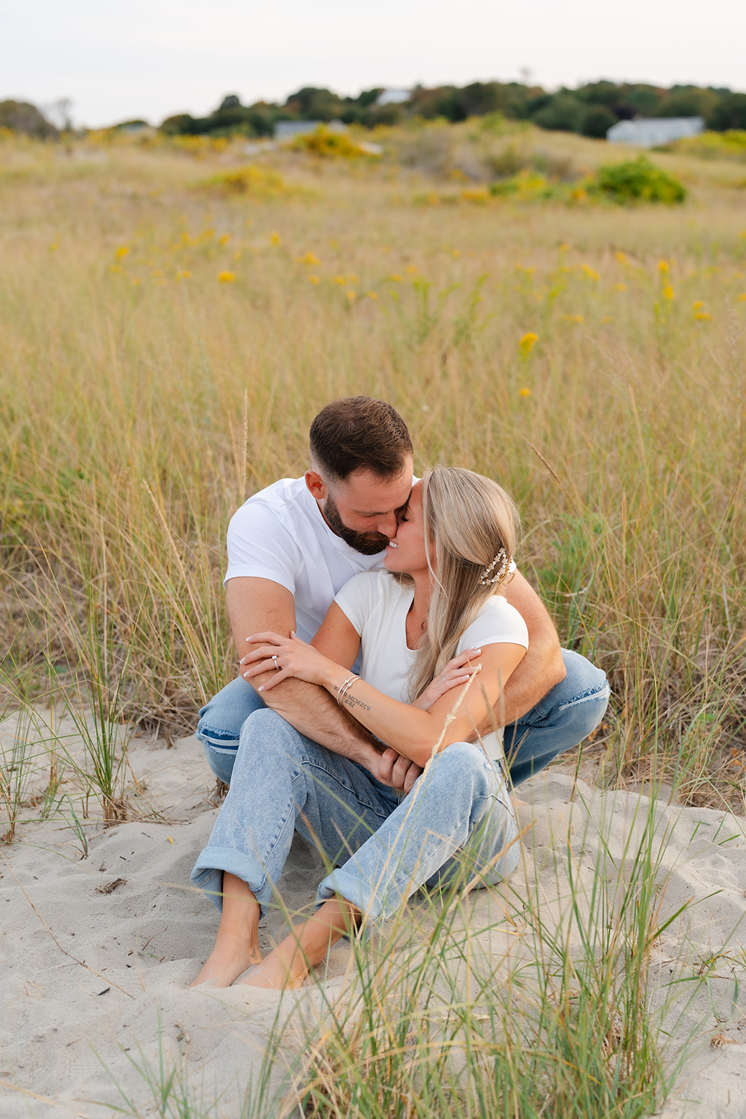 A couple sharing a tender moment during their beach engagement session, captured against the soft light and greenery around them.