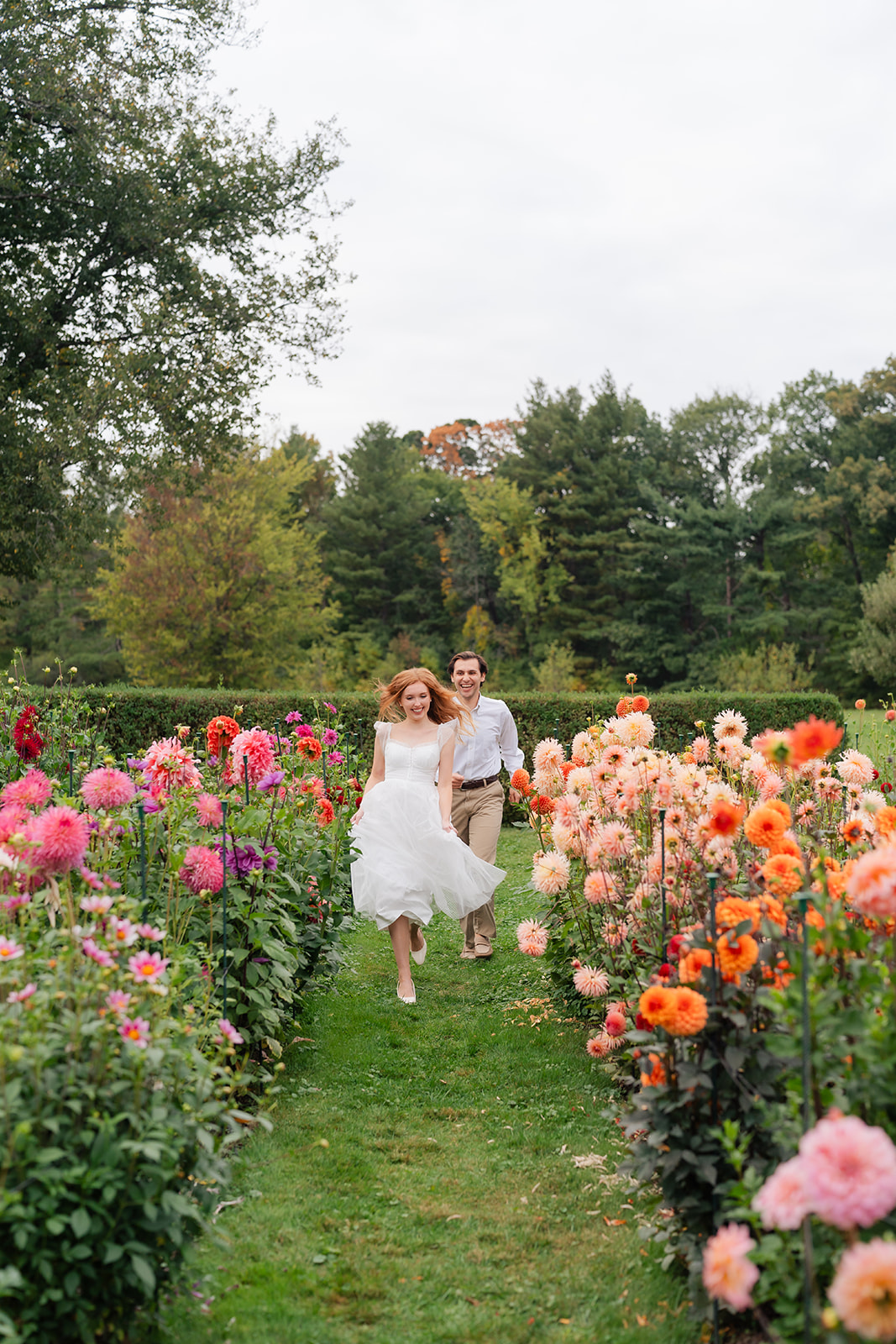 A couple running surrounded by blooming flowers in a picturesque garden, showcasing their love and unique engagement shoot ideas at Stevens-Coolidge House & Gardens.