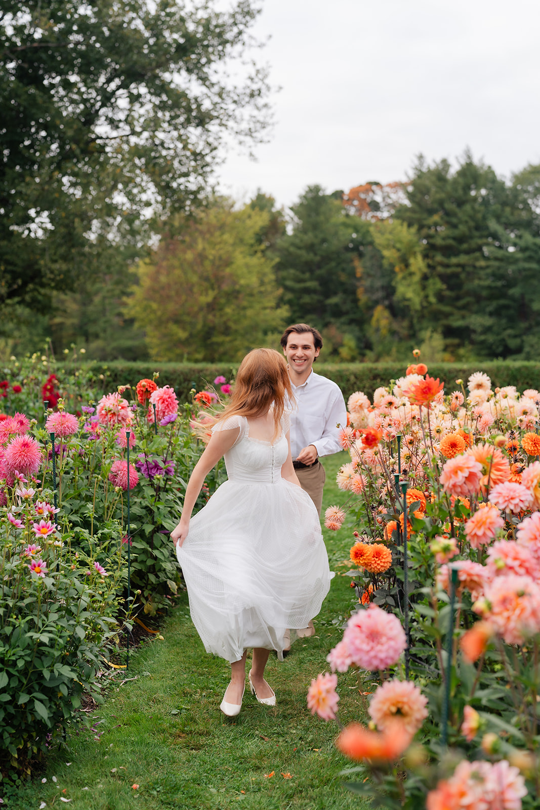 A couple running surrounded by blooming flowers in a picturesque garden, showcasing their love and unique engagement shoot ideas at Stevens-Coolidge House & Gardens.