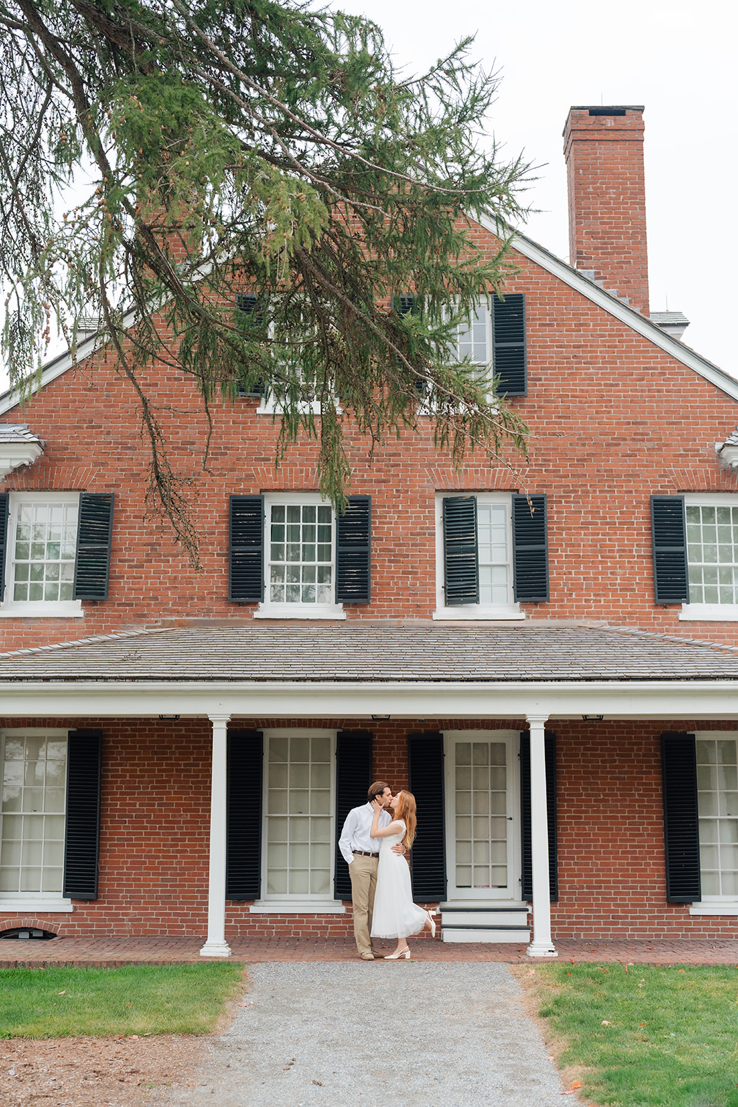 The couple sharing a sweet kiss outside a brick house with a large tree overhead, creating a timeless and classic engagement photo. The simplicity of the location highlights their connection, making it one of those unique engagement shoot ideas that never goes out of style.