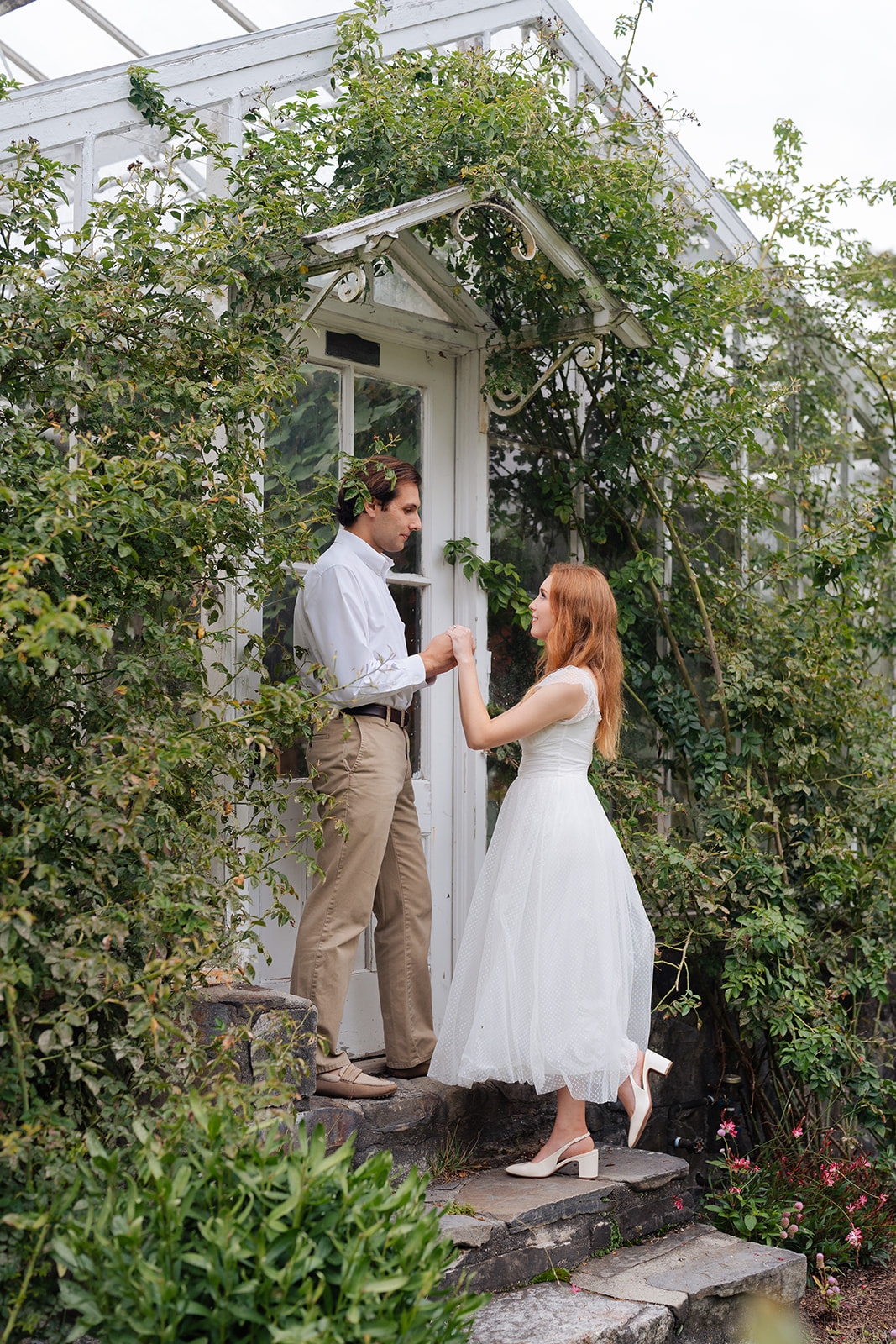 The couple holding hands and laughing, with flowers and plants surrounding them in a whimsical garden setting. A perfect example of unique engagement shoot ideas, this image exudes joy and nature's beauty, adding a fresh and lively vibe to the photos.