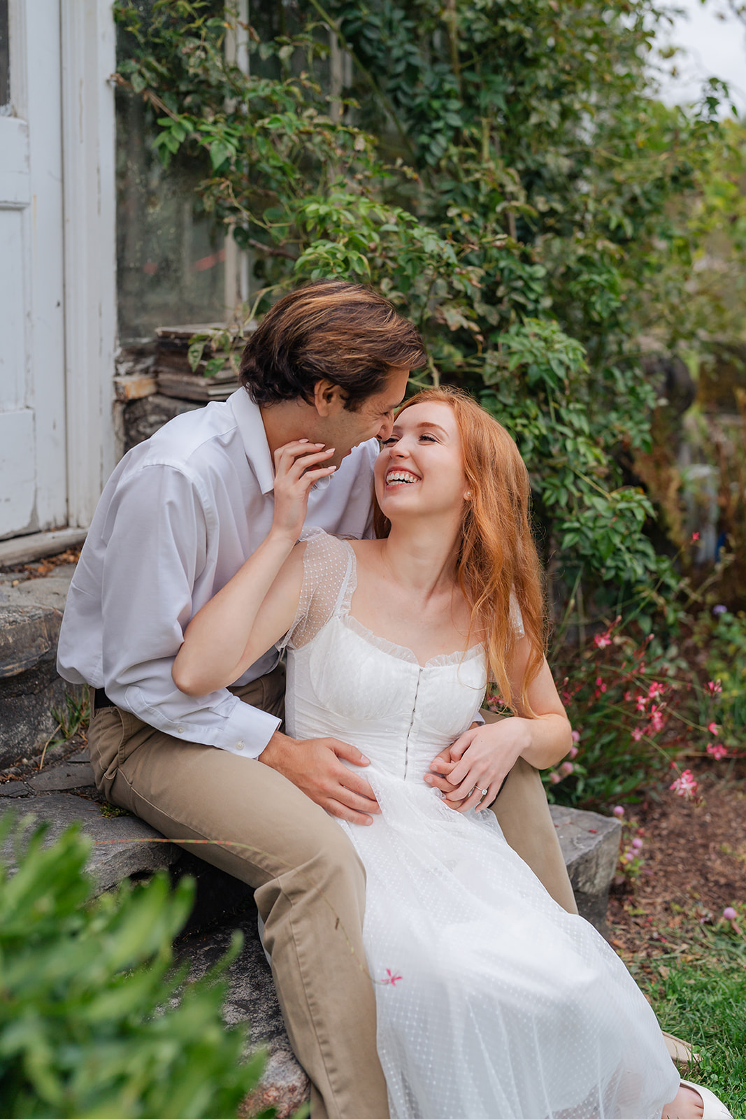 The couple holding hands and laughing, with flowers and plants surrounding them in a whimsical garden setting. A perfect example of unique engagement shoot ideas, this image exudes joy and nature's beauty, adding a fresh and lively vibe to the photos.